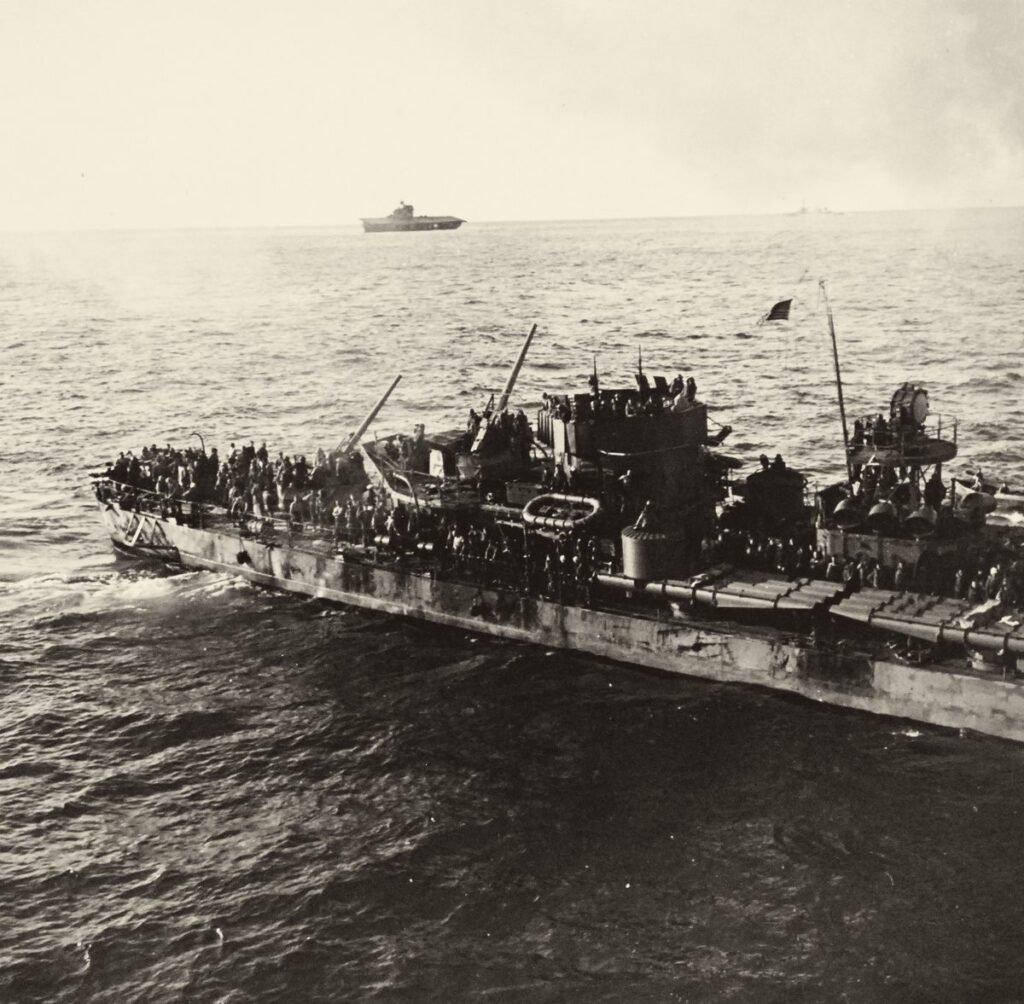 Vista do alto: um navio de guerra estreito, com o convés lotado de homens; ao longe, no horizonte, um porta-aviões.